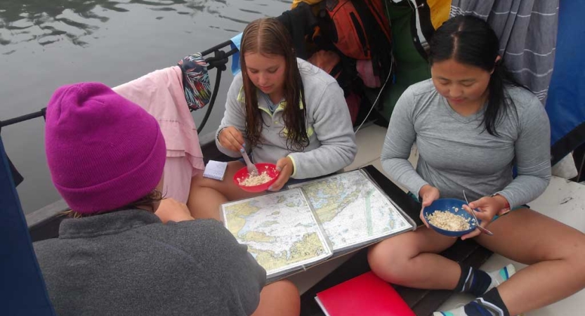 Three students sitting in a sailboat eat while they examine a map.